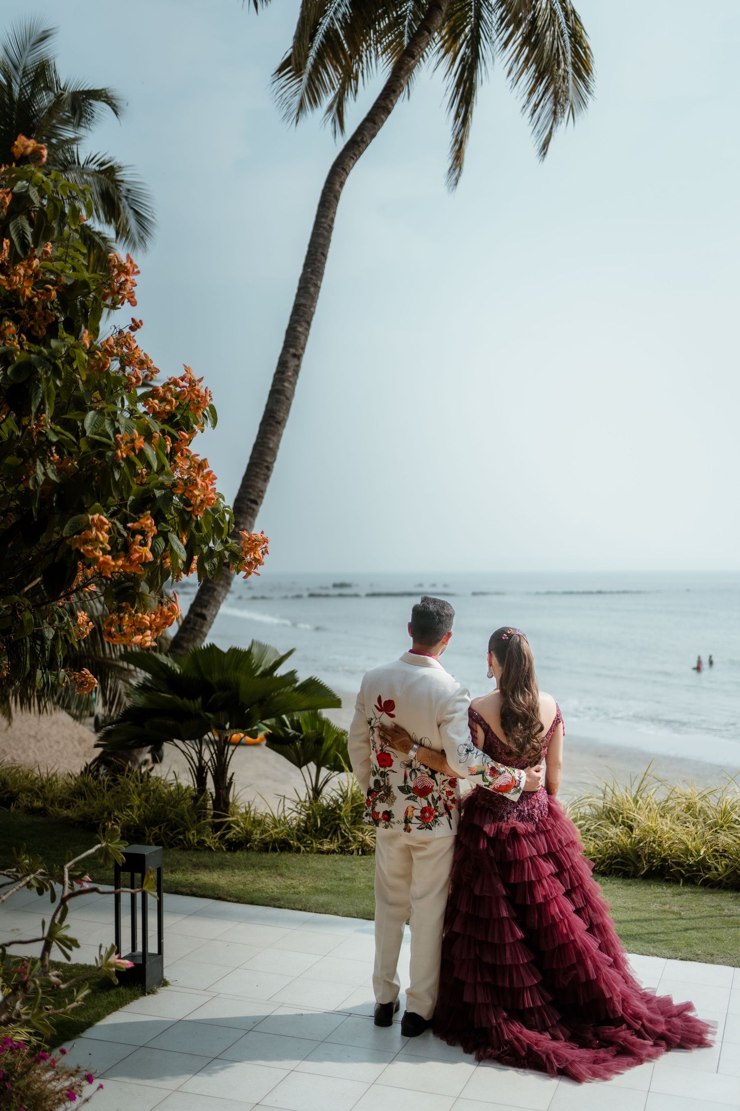 A couple in wedding attire stands on a beachfront terrace with palm trees and ocean views in the background.