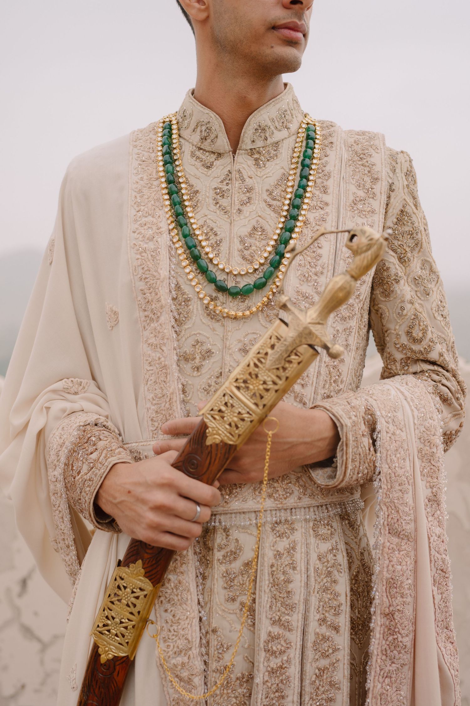 Traditional Indian groom wearing cream sherwani with intricate embroidery and layered pearl necklace holding ceremonial sword.