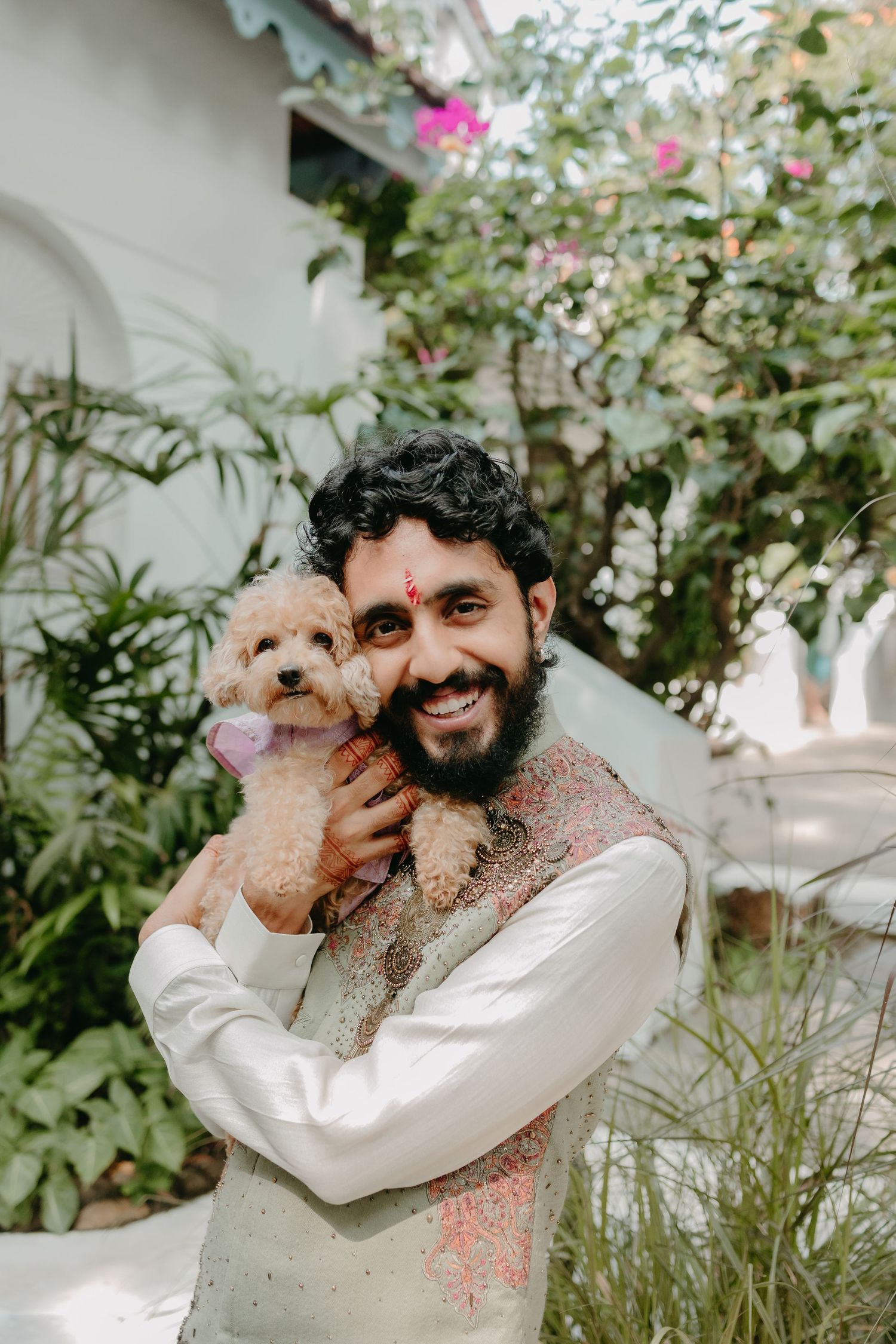 Smiling bearded Indian man in traditional kurta holding a fluffy apricot poodle puppy outdoors during Holi festival.