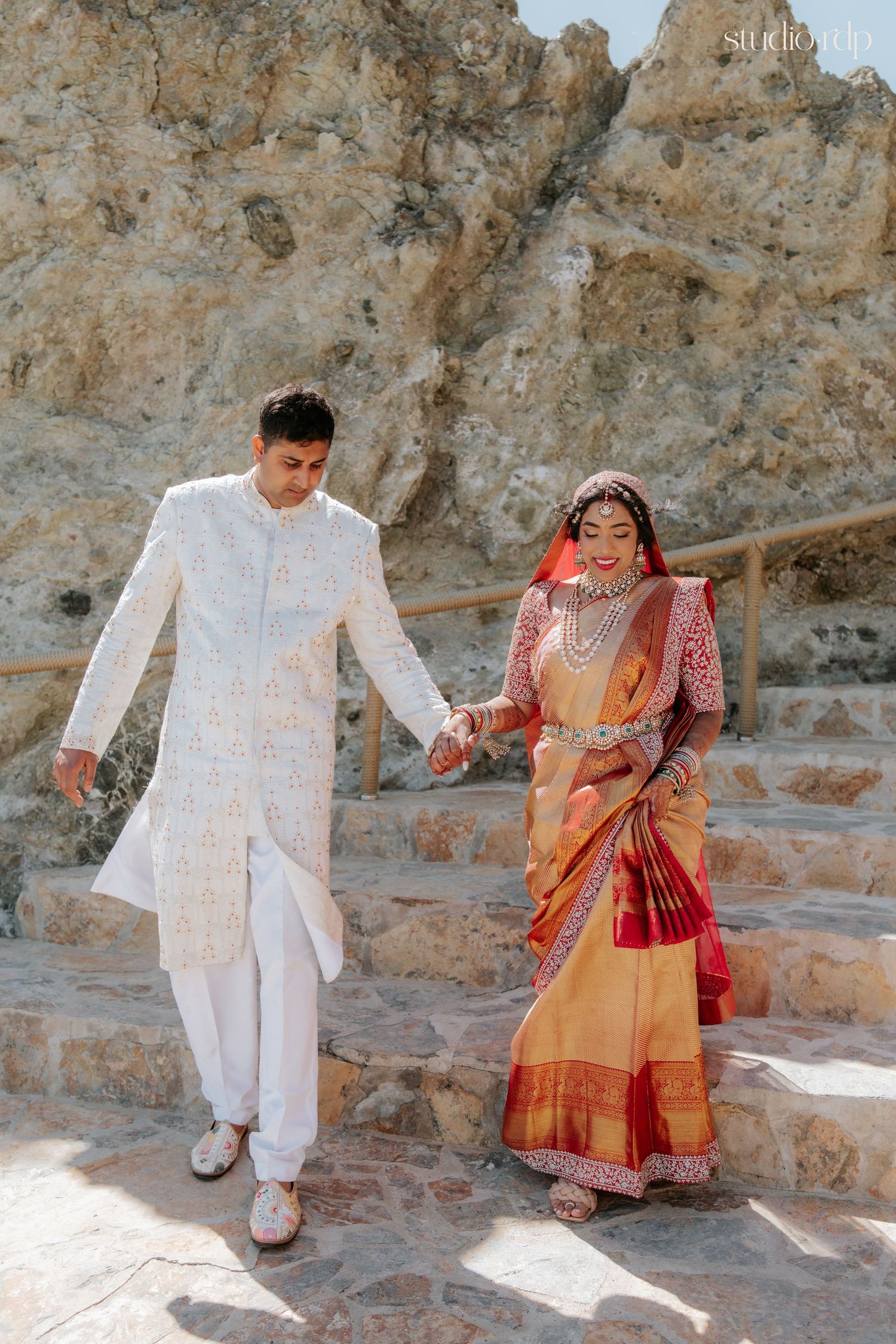Indian couple in traditional wedding attire walking down stone steps, groom in white sherwani, bride in orange saree.
