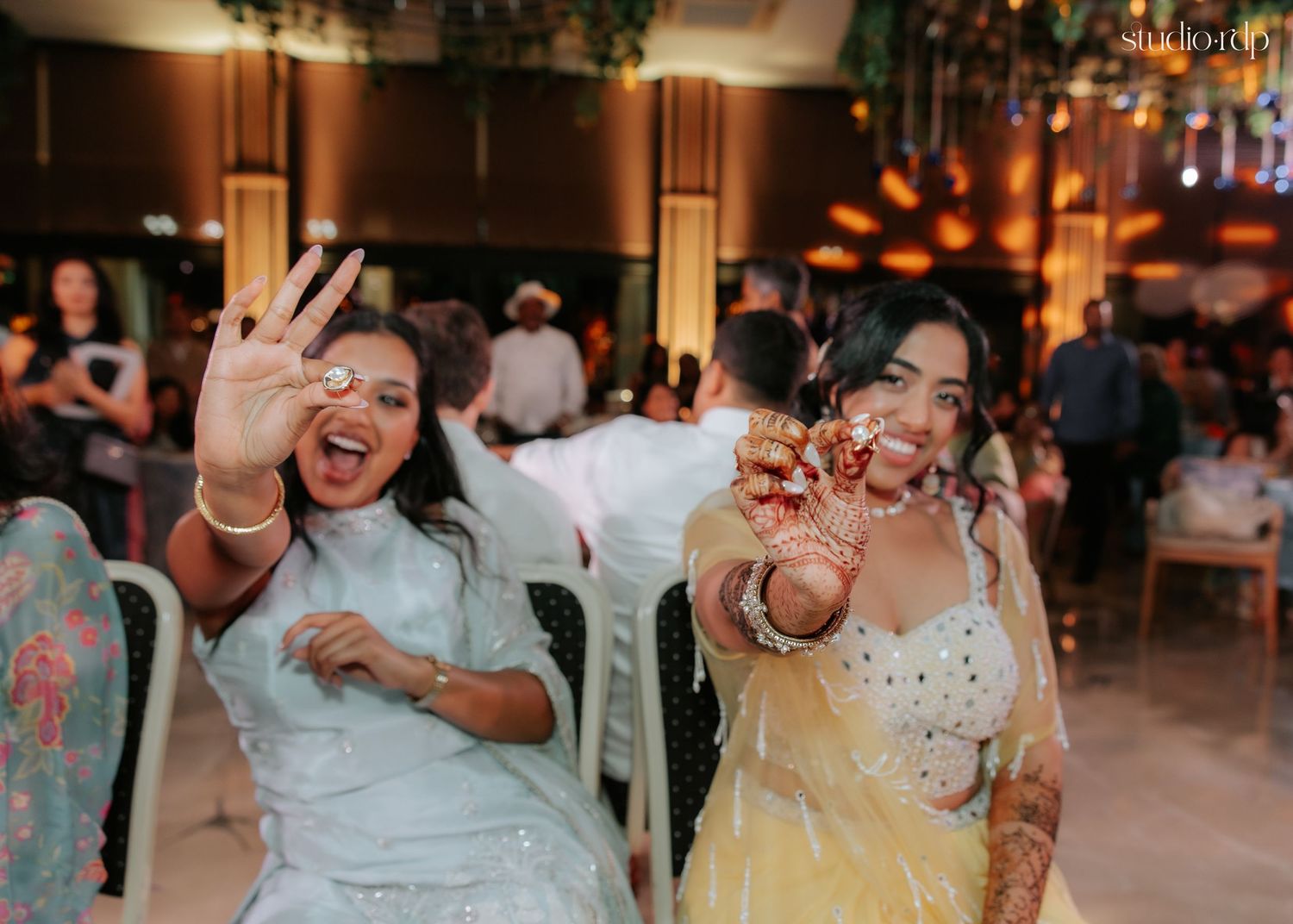 Two women in colorful traditional attire smiling and holding sparklers at a festive Indian wedding celebration.