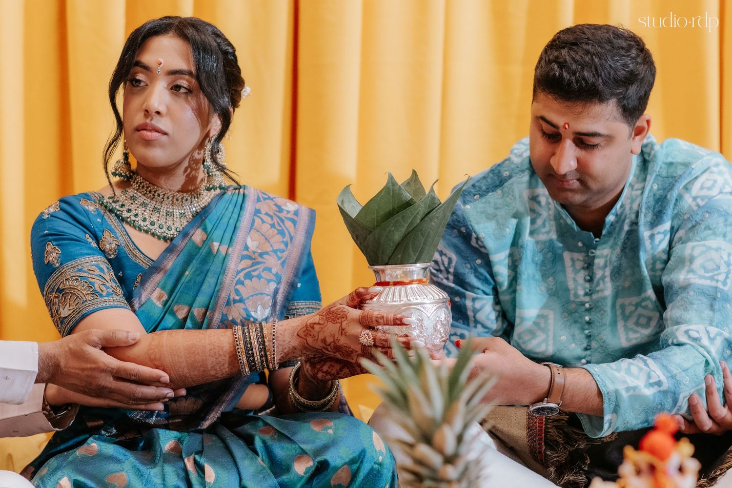 Indian couple in traditional blue attire performing a wedding ritual with a decorated pot during a ceremony.