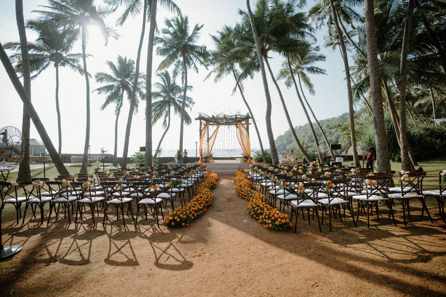 Outdoor beach wedding ceremony setup with rows of chairs, floral aisle, wooden arch, and tall palm trees in tropical setting.