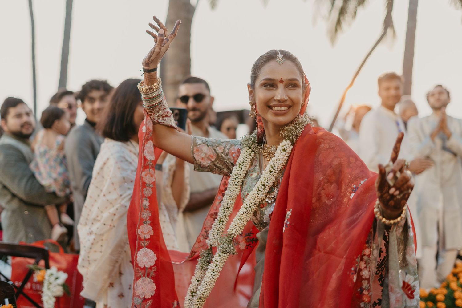 Smiling bride in red saree with floral garland dancing at outdoor Indian wedding ceremony with guests in background.