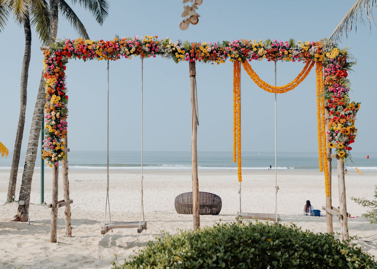 A beachside swing decorated with colorful flowers and yellow fabric draping under a floral arch by the ocean.