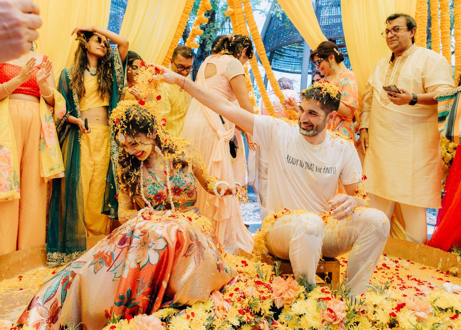 Flower petals shower guests at a colorful Indian wedding ceremony with people gathered in traditional attire.