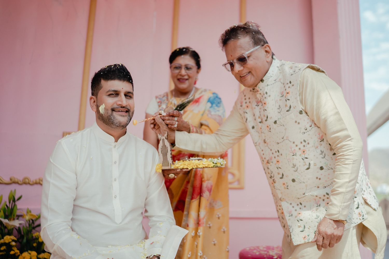 Three people share a joyful moment during an Indian wedding ceremony, wearing traditional attire against a pink backdrop.