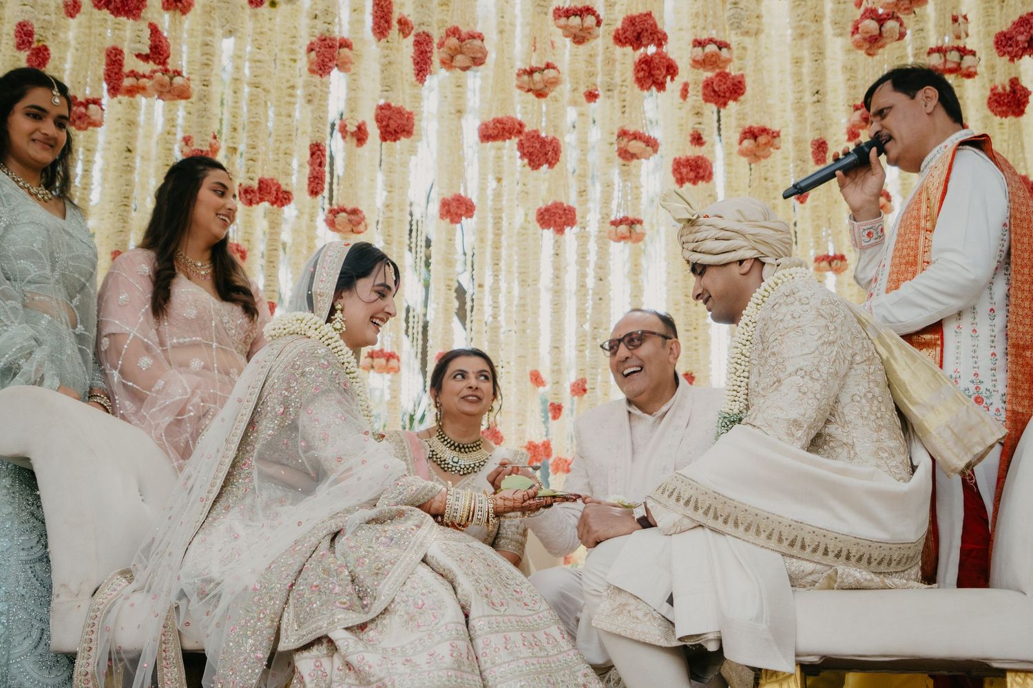 Traditional Indian wedding ceremony with participants seated on white furniture against a backdrop of red floral decorations.