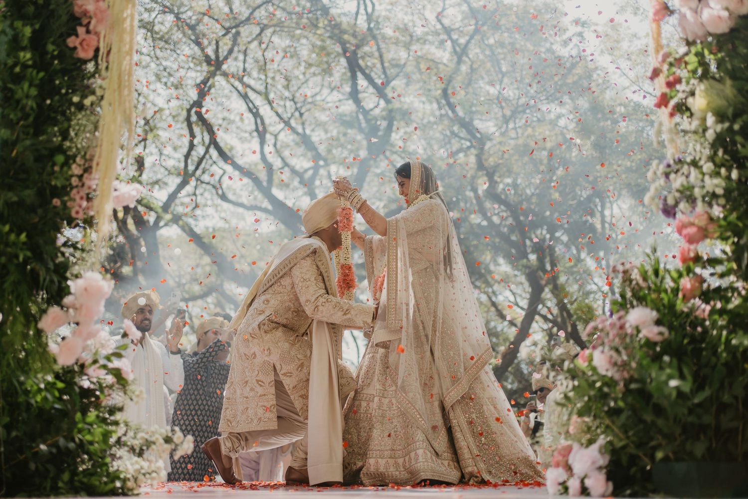 A couple in traditional wedding attire embraces under flowering cherry blossom trees in a romantic outdoor garden setting.
