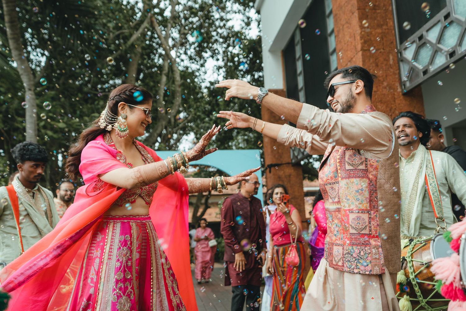 Bride and groom in vibrant traditional Indian wedding attire dancing joyfully outdoors with musicians and guests.