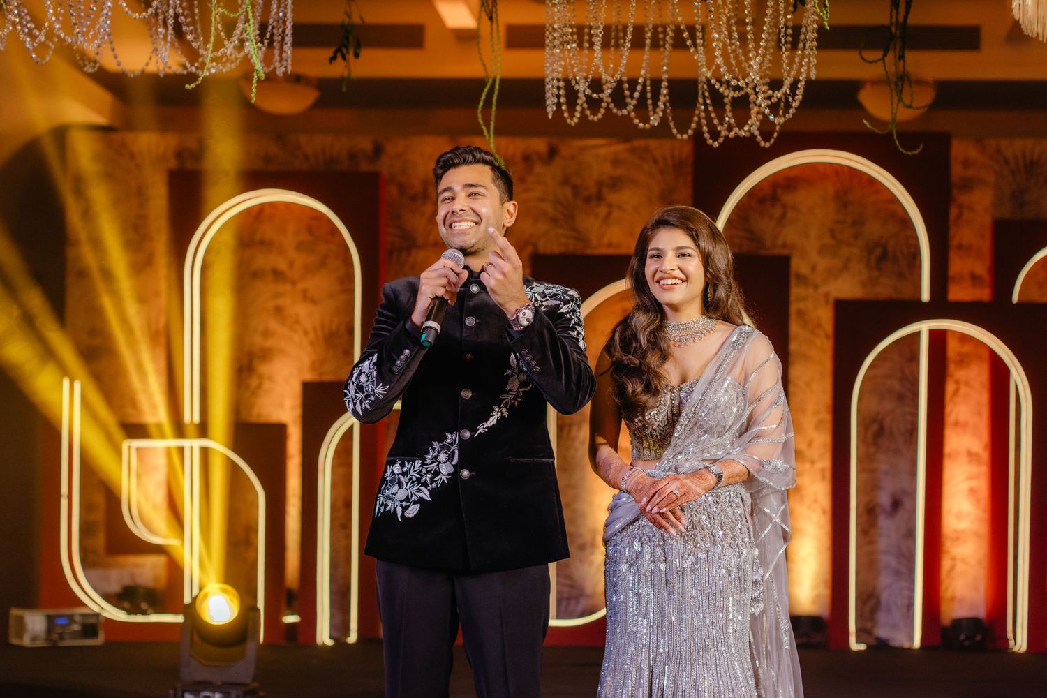 Two performers sing together on stage with illuminated arches and chandeliers in the warm-lit background.