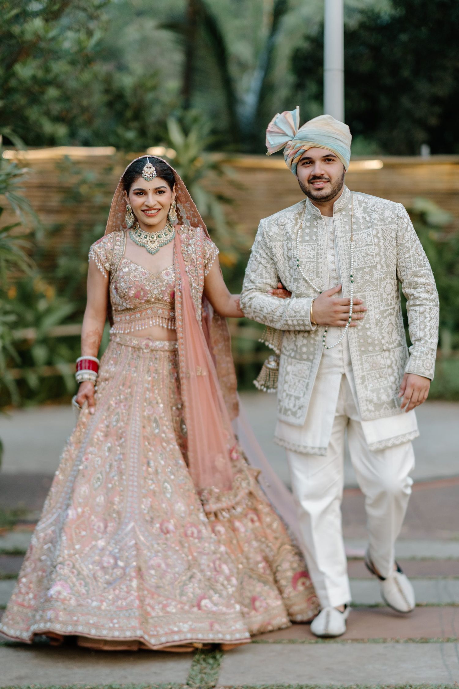 A couple in traditional Indian wedding attire with the bride in pink lehenga and groom in white sherwani pose outdoors.