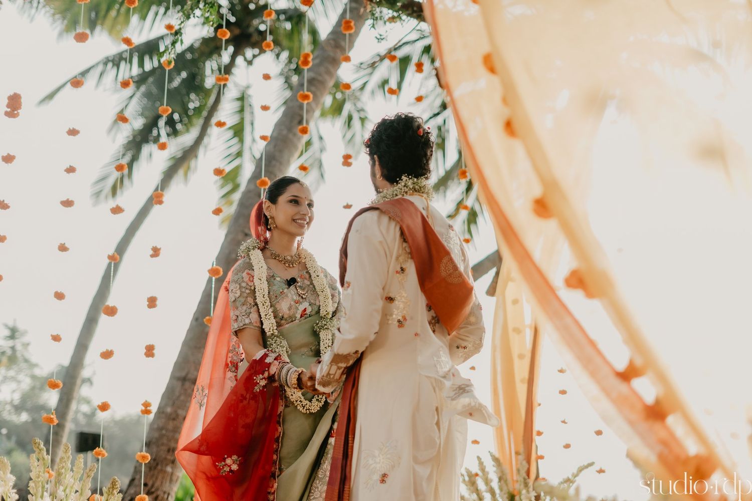 Couple in traditional Indian wedding attire standing under a decorated canopy with orange drapes and string lights.