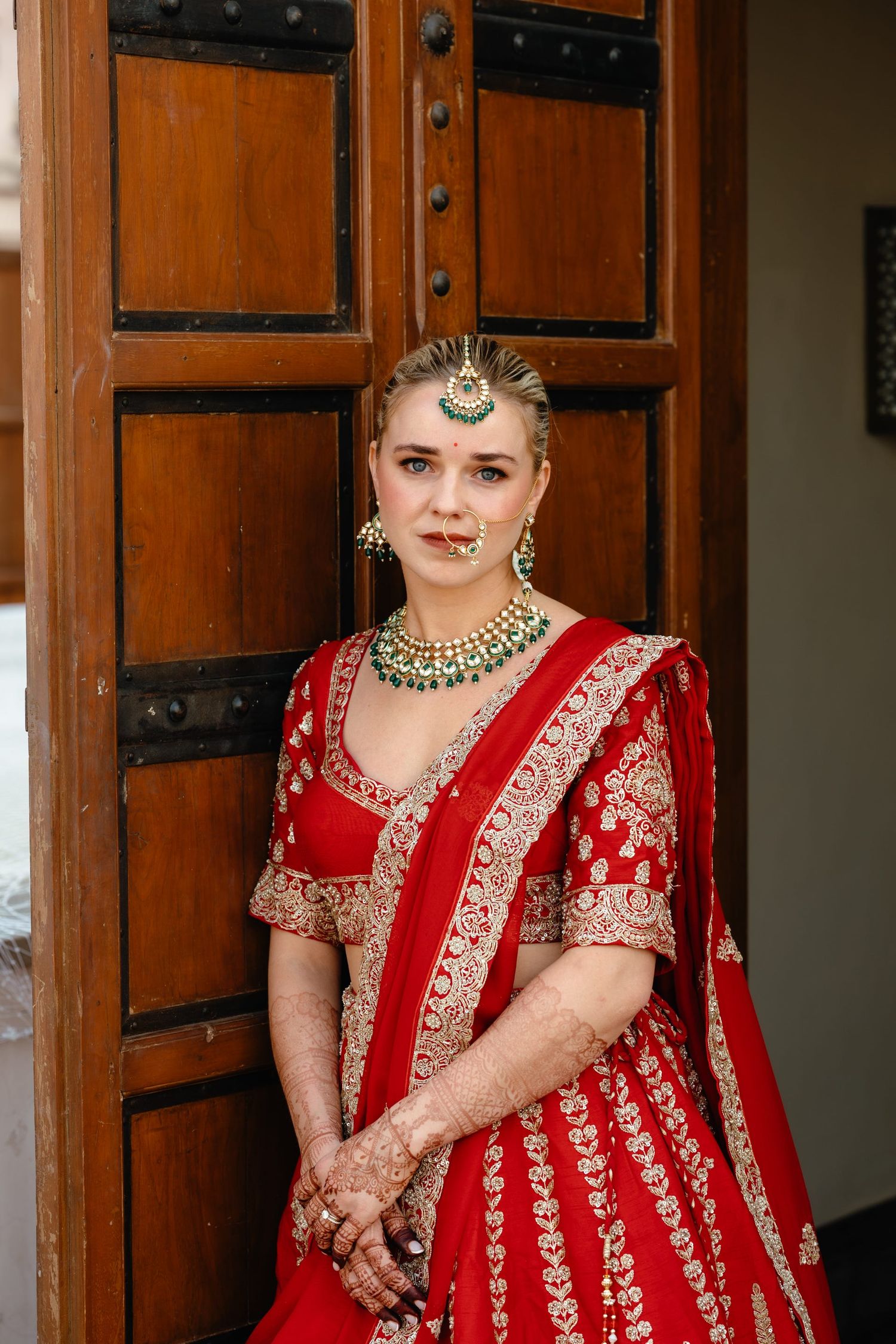 Traditional red bridal outfit with intricate gold embroidery photographed against wooden doors.