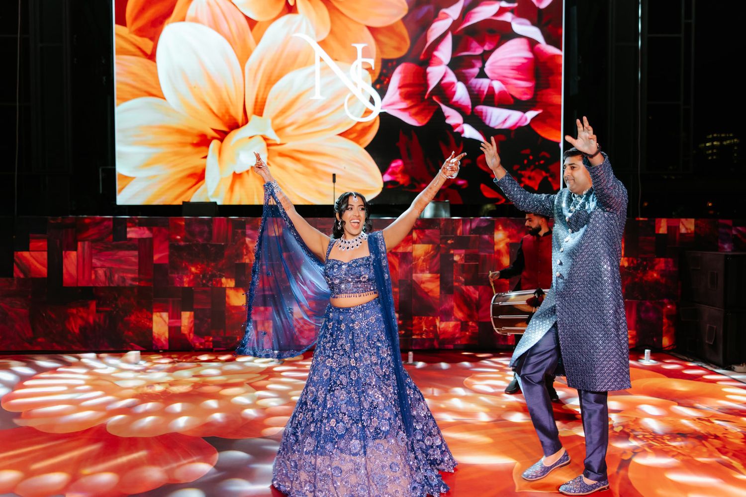 A couple in blue traditional Indian outfits dance joyfully on stage with a floral LED display backdrop.