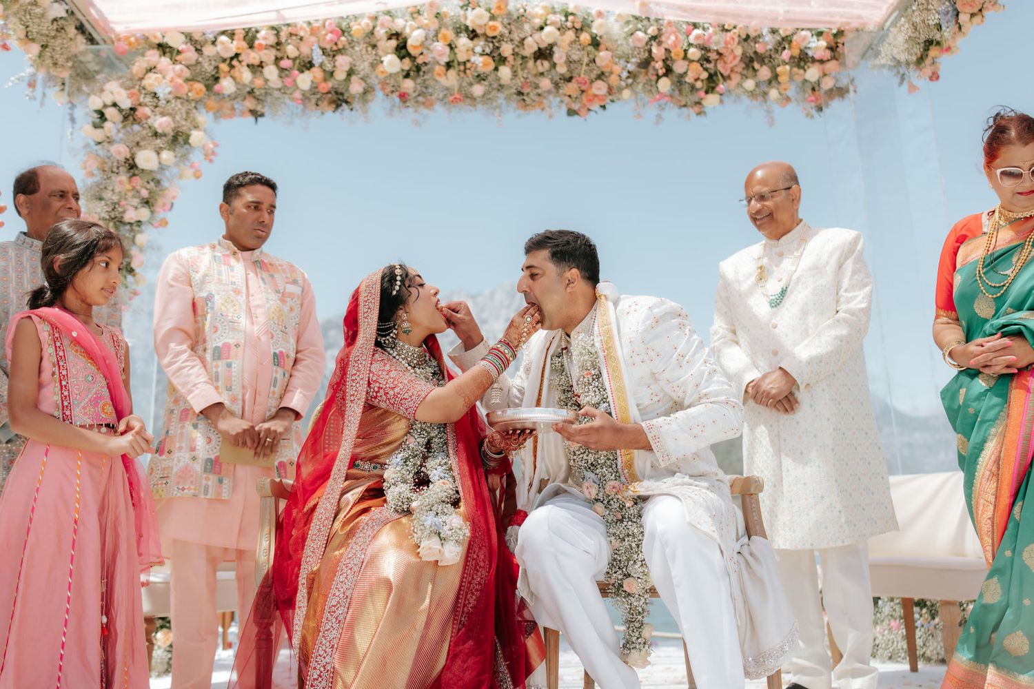 Indian bride and groom exchange garlands during traditional Hindu wedding ceremony outdoors under floral mandap with guests.