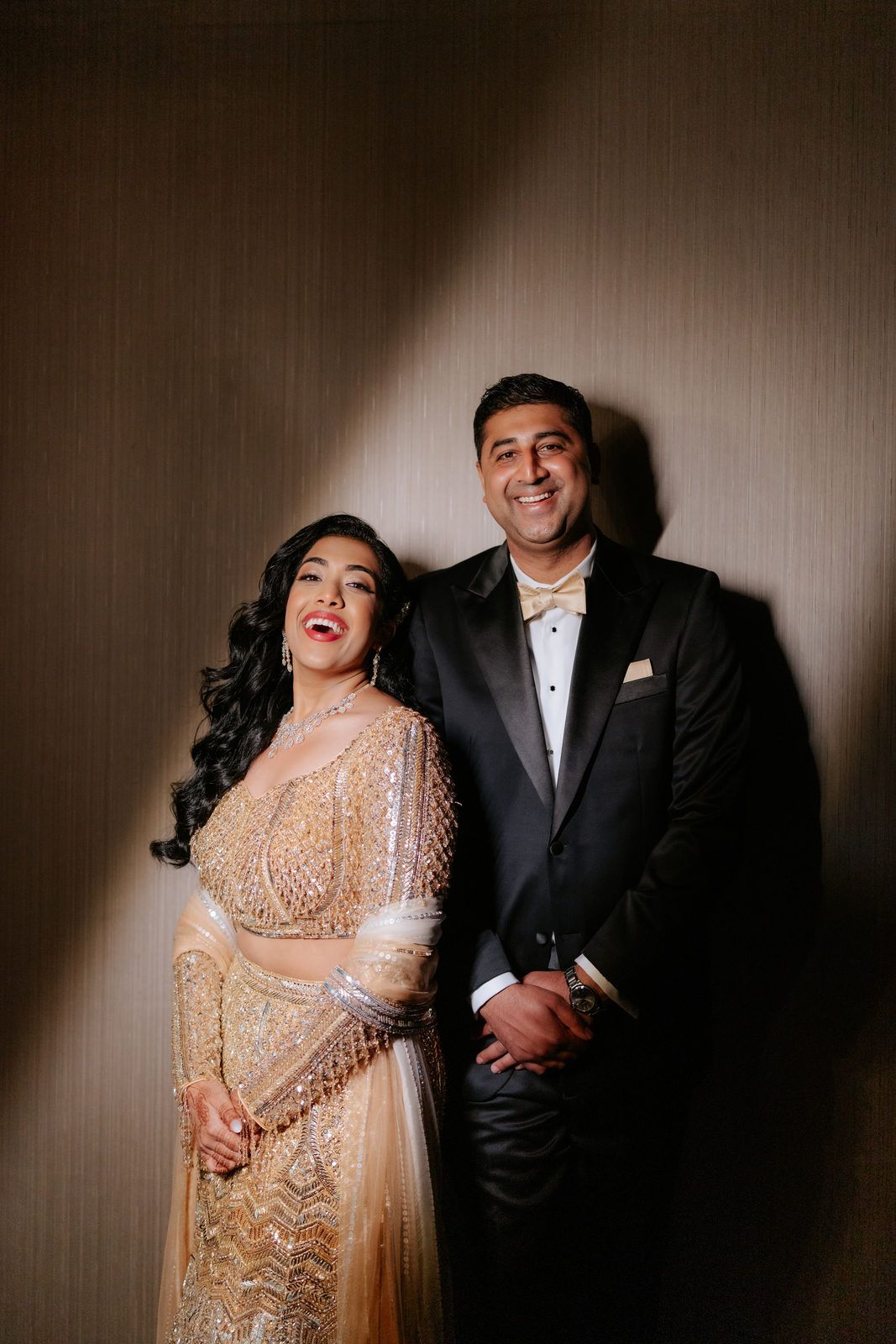 Joyful Indian wedding couple laughing together, bride in gold beaded lehenga and groom in black tuxedo.