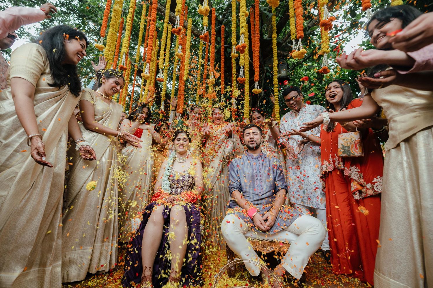 Joyful Indian wedding couple showered with flower petals by guests under colorful floral decorations outdoors.