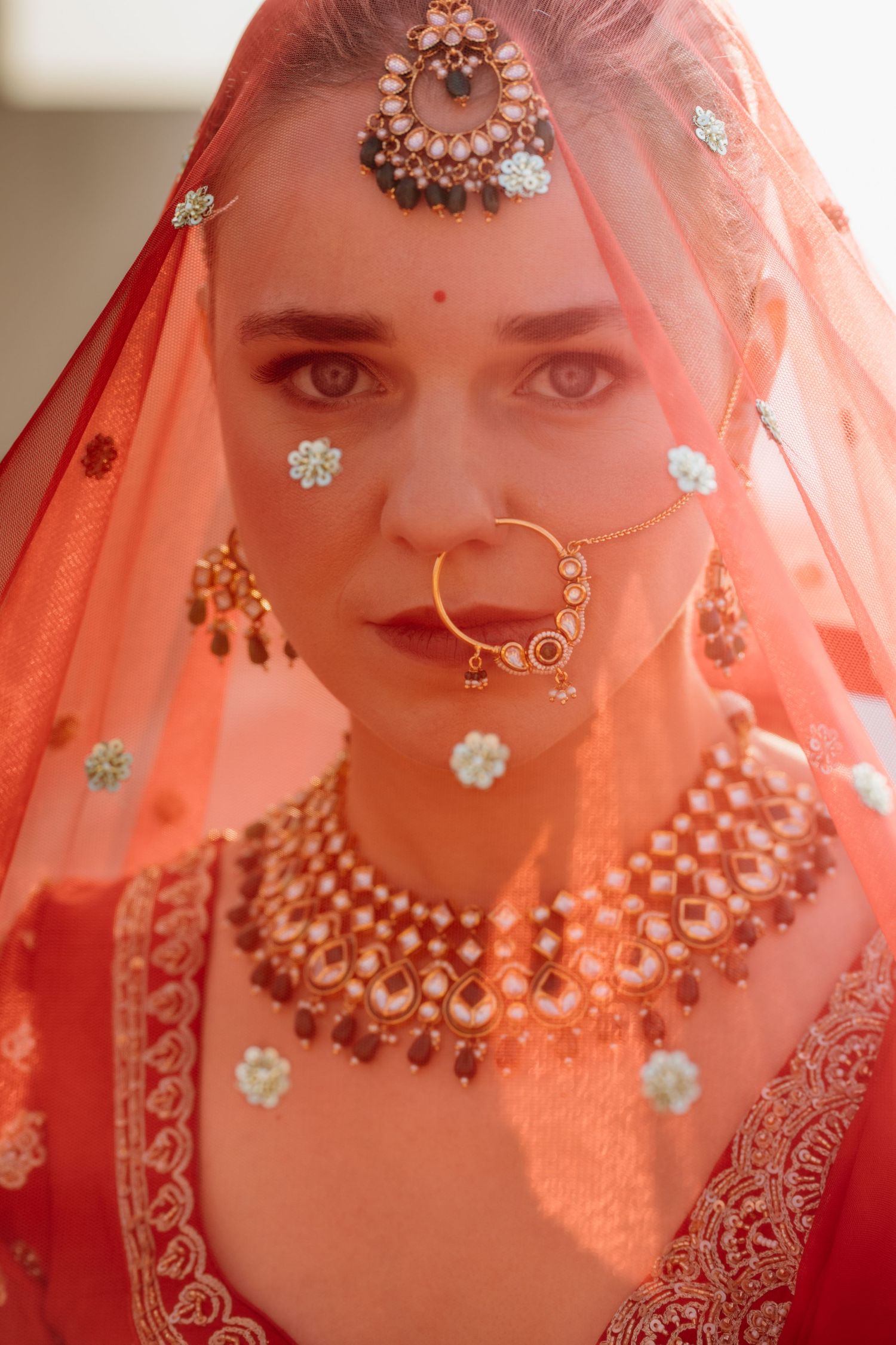 A bride in red lehenga with gold kundan jewelry, maang tikka, nose ring, and red dupatta veil.