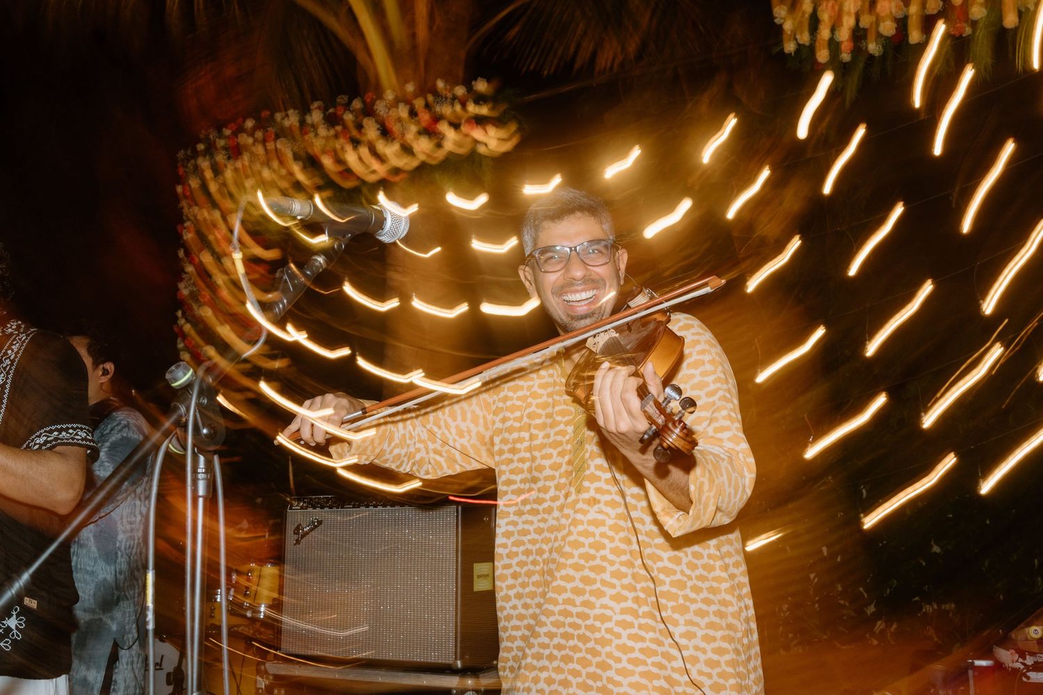 Man in traditional attire playing violin at night with glowing light trails swirling in the background.