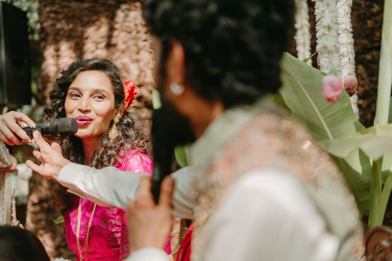Woman in pink saree singing into microphone at colorful Indian wedding ceremony outdoors.