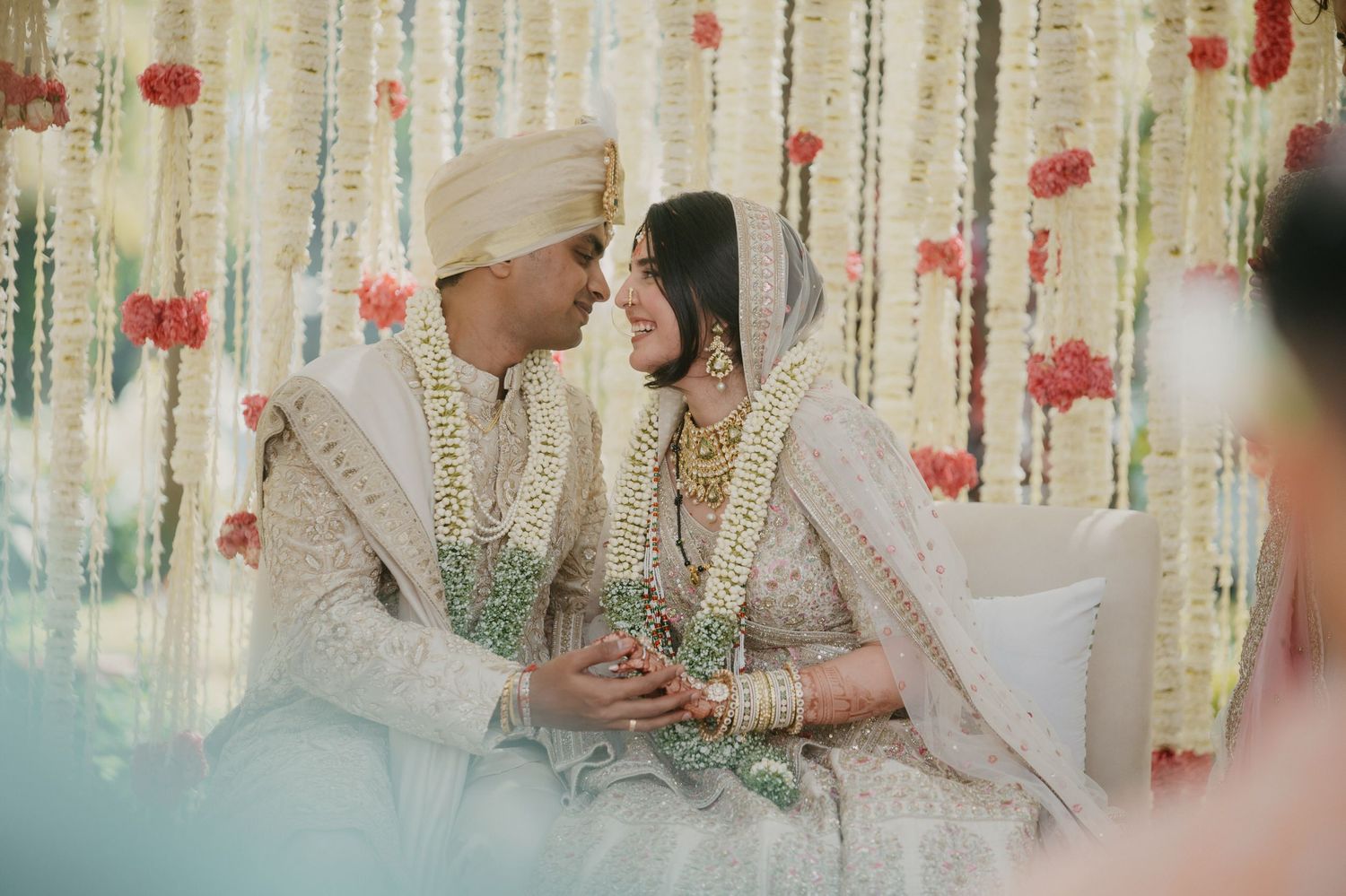 A couple in traditional white and gold Indian wedding attire sit together under elegant floral decorations at their ceremony.