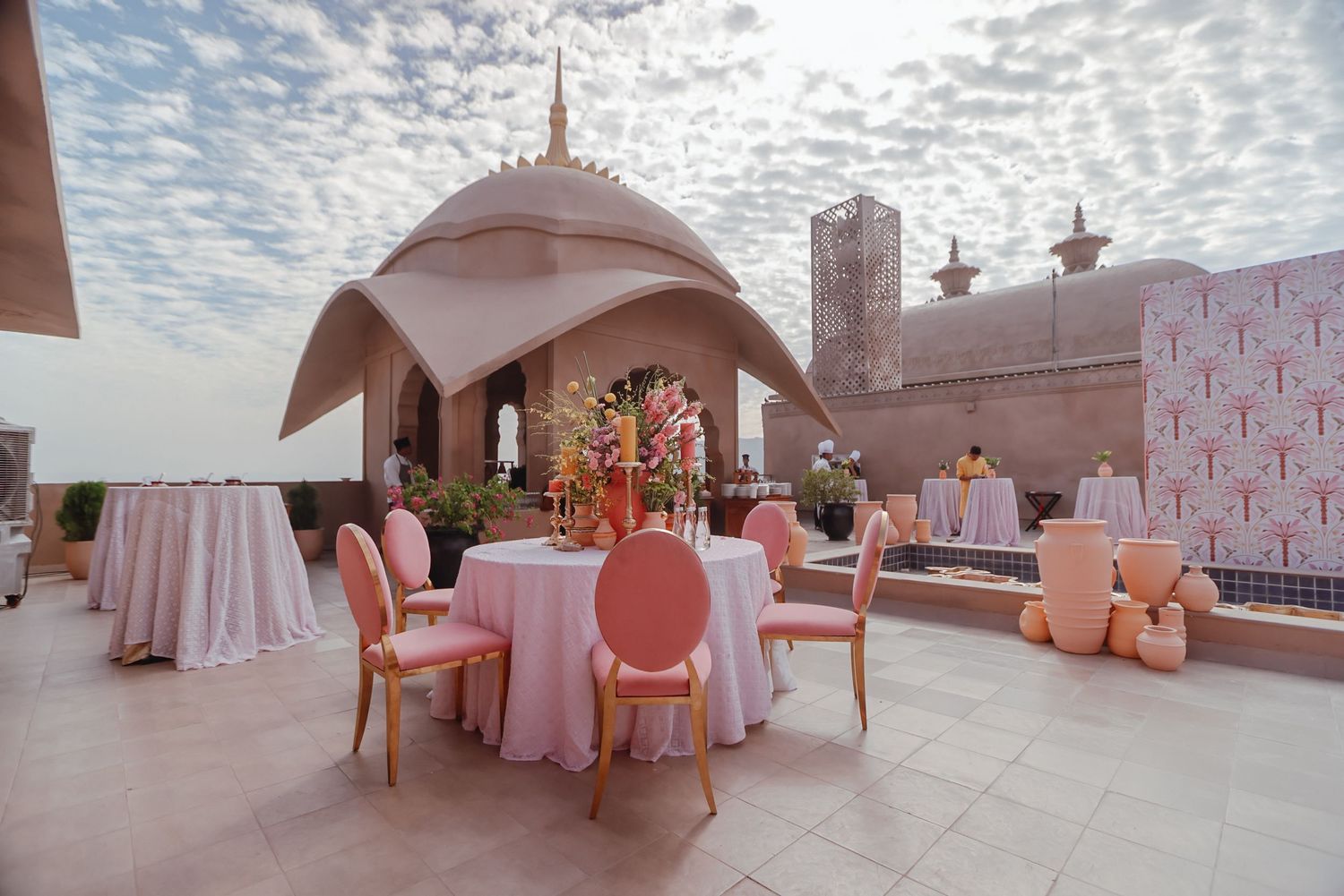 Pink and coral rooftop wedding reception setup with elegant tables under an arched canopy against a cloudy sunset sky.