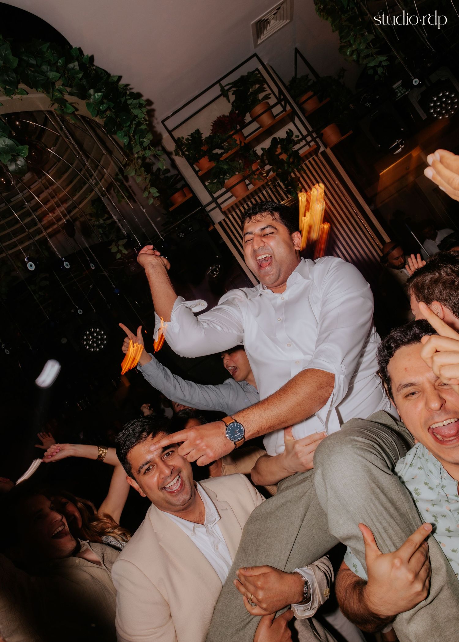 Men celebrating joyfully at a party, one lifted on shoulders, smiling and dancing in a festive indoor venue.