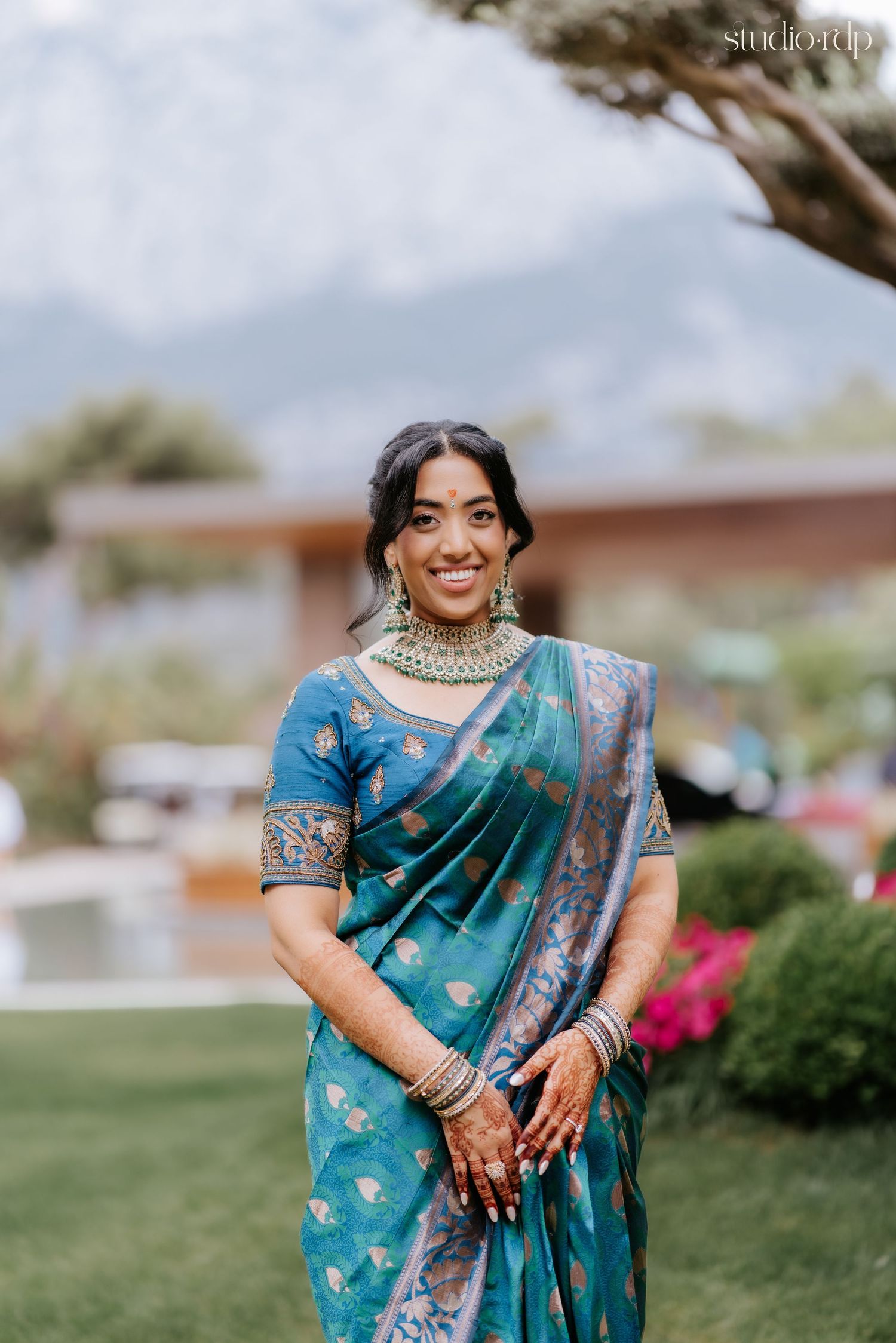 Smiling bride in teal silk saree with gold jewelry and mehndi, posing outdoors with mountains in background.