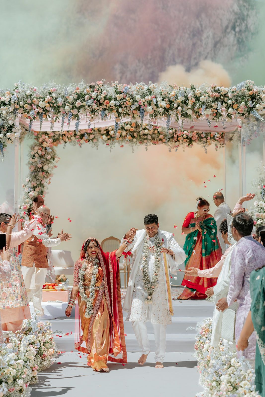 Indian bride and groom exit wedding mandap under colorful smoke with floral arch and celebrating guests.