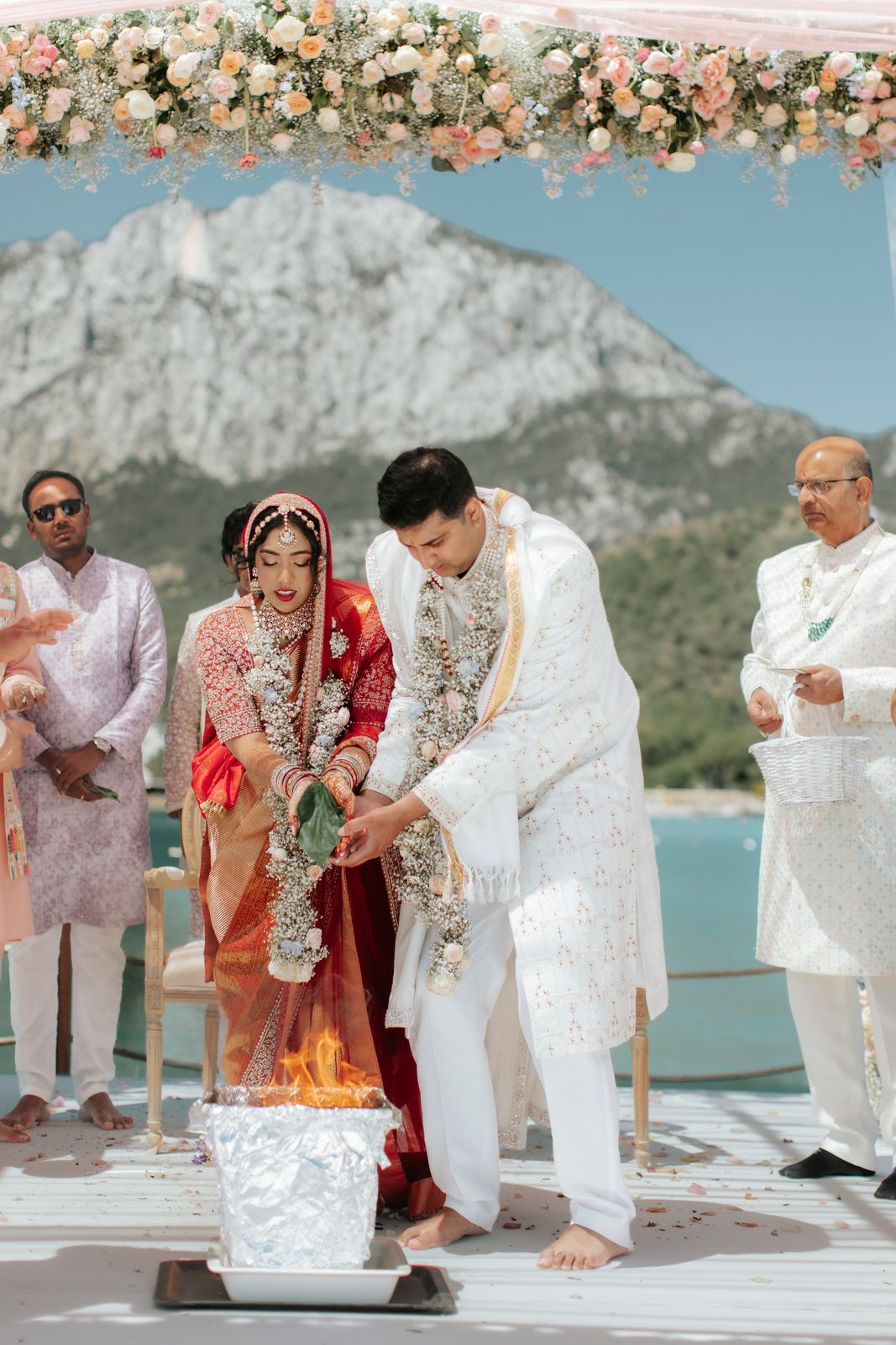 Indian couple performing sacred fire ritual at outdoor Hindu wedding ceremony with mountain and ocean backdrop in white attire.