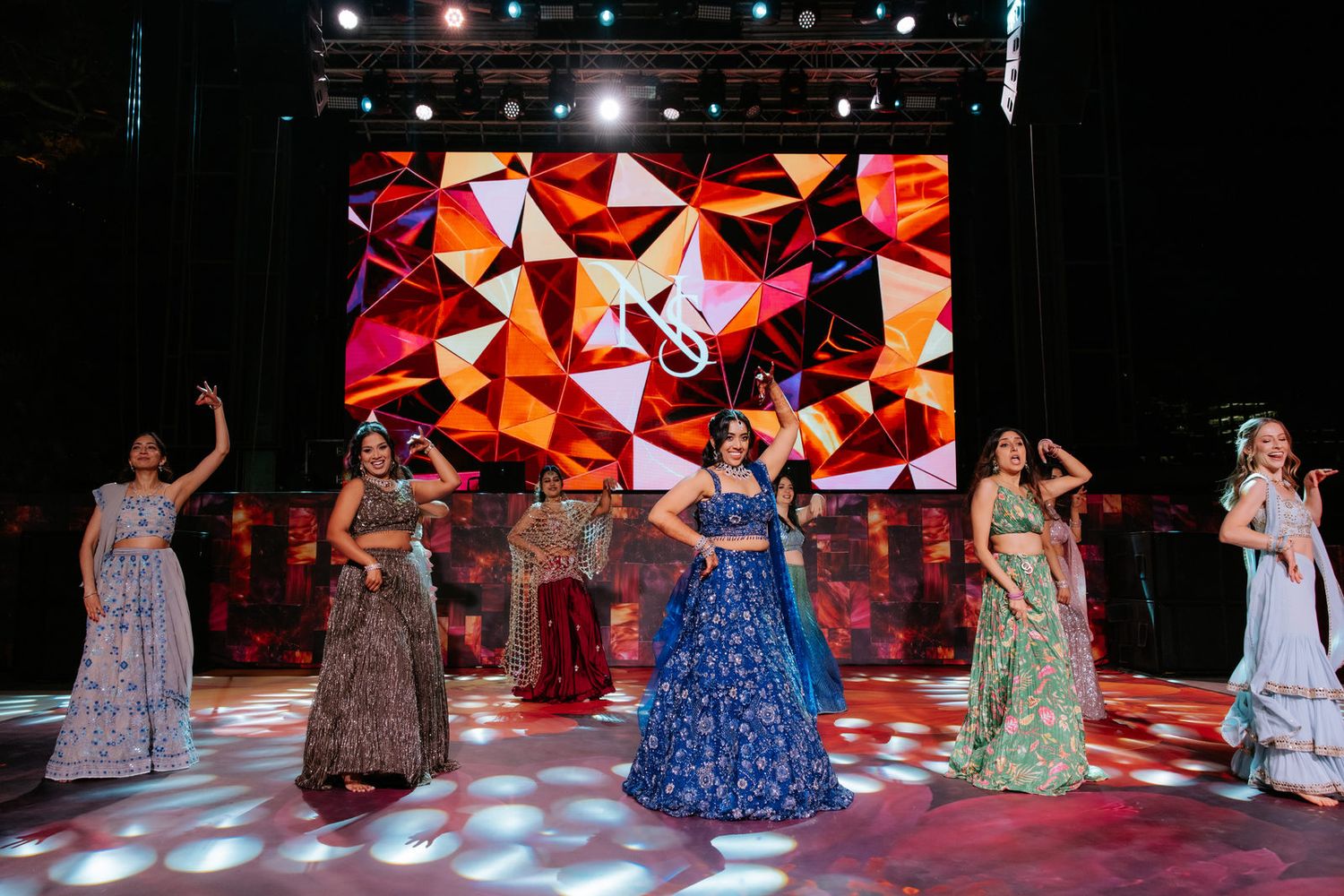 Group of women in colorful lehengas performing on stage with vibrant geometric LED backdrop.