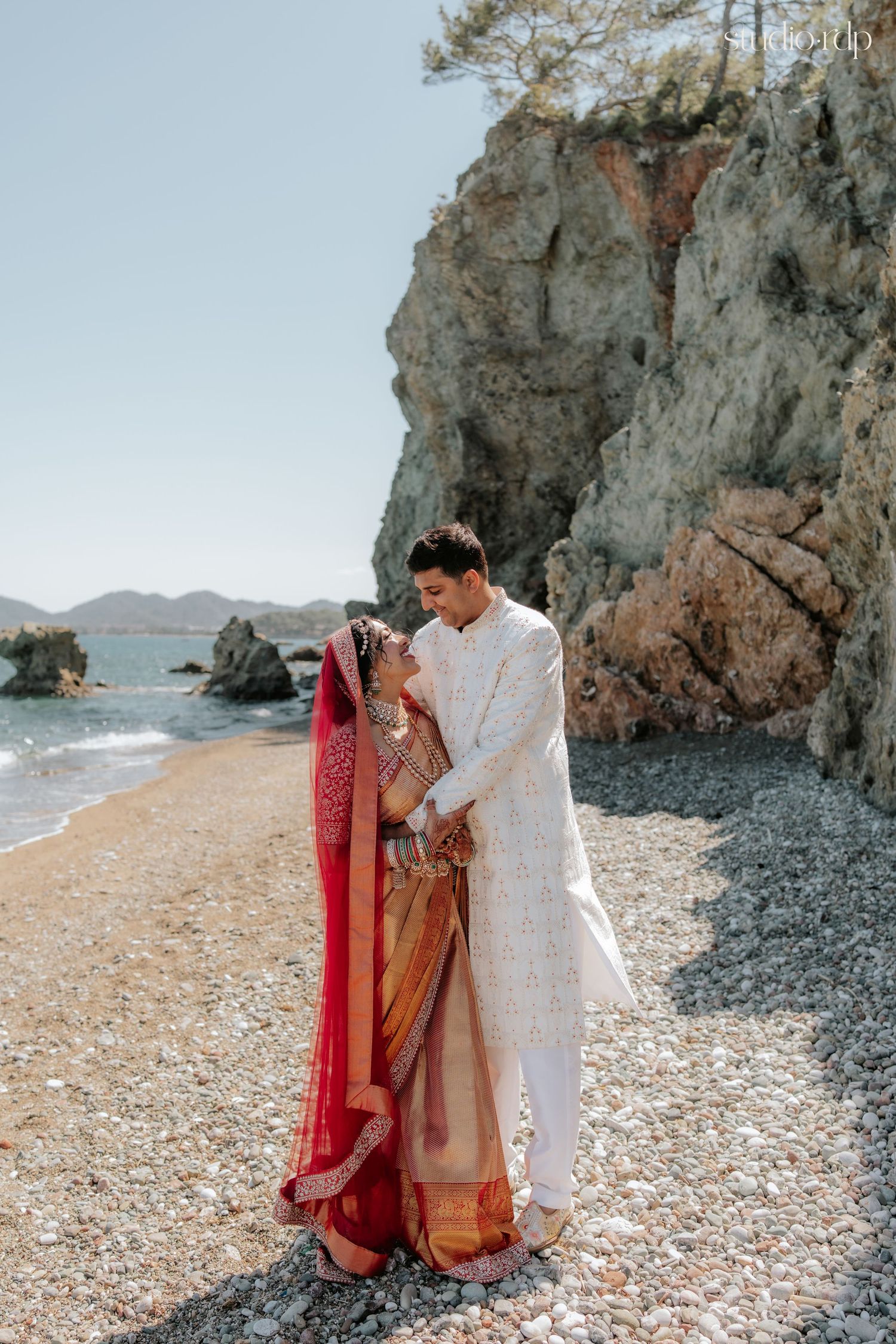 Indian couple in traditional wedding attire sharing a romantic moment on a rocky beach shoreline.