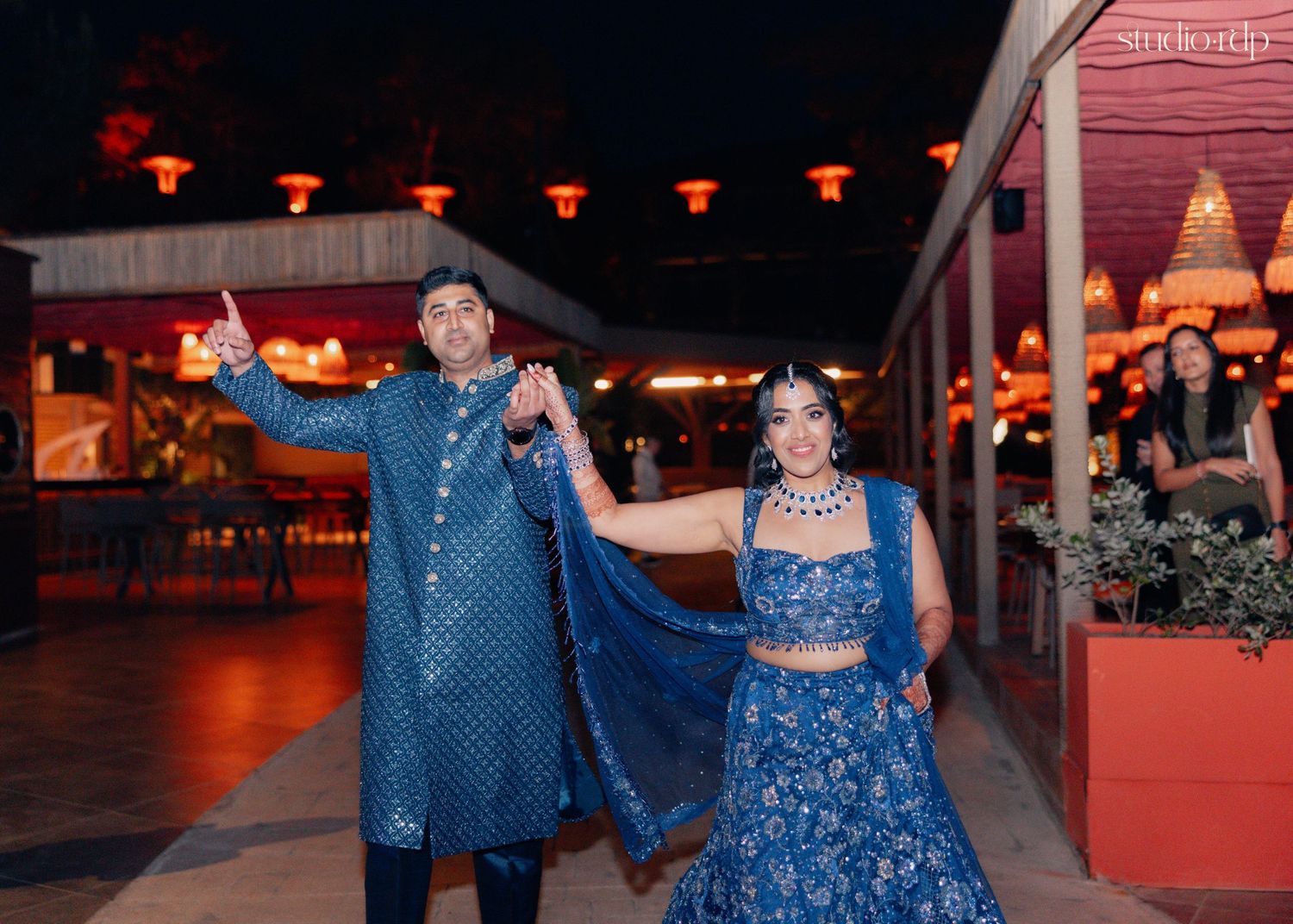Couple in matching blue Indian wedding attire dancing at an outdoor evening celebration venue.
