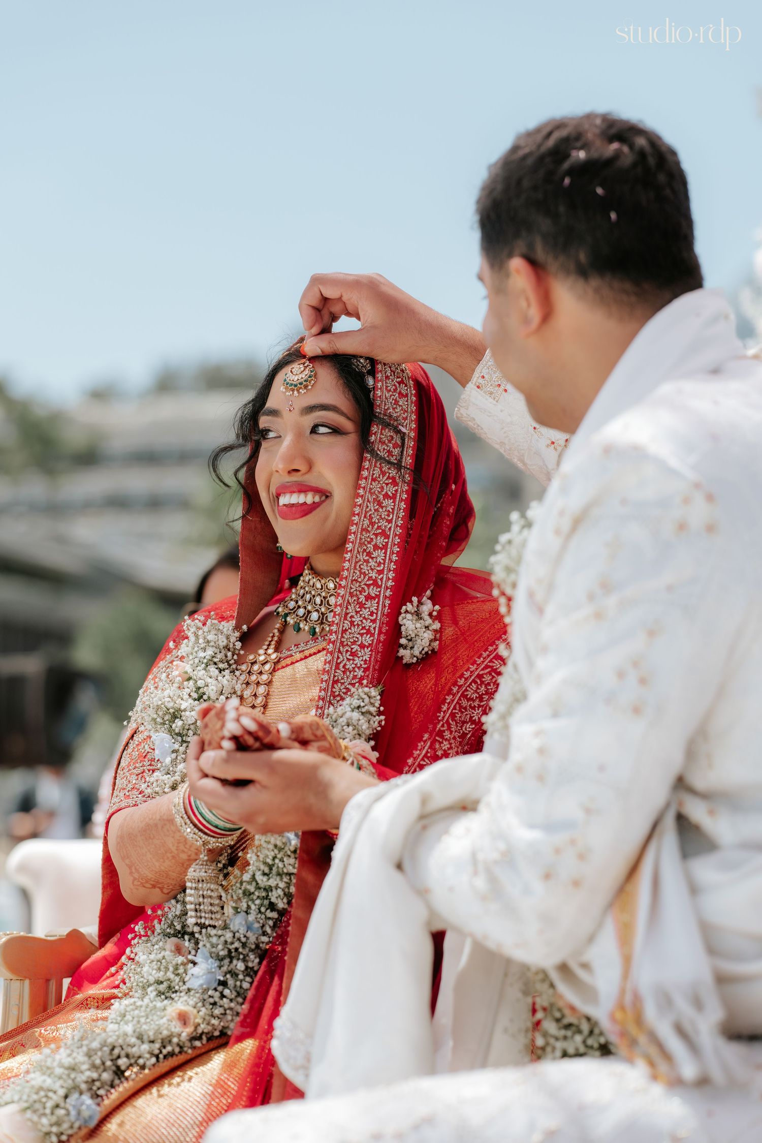 Groom applying sindoor to smiling bride in red lehenga during traditional Indian wedding ceremony outdoors.