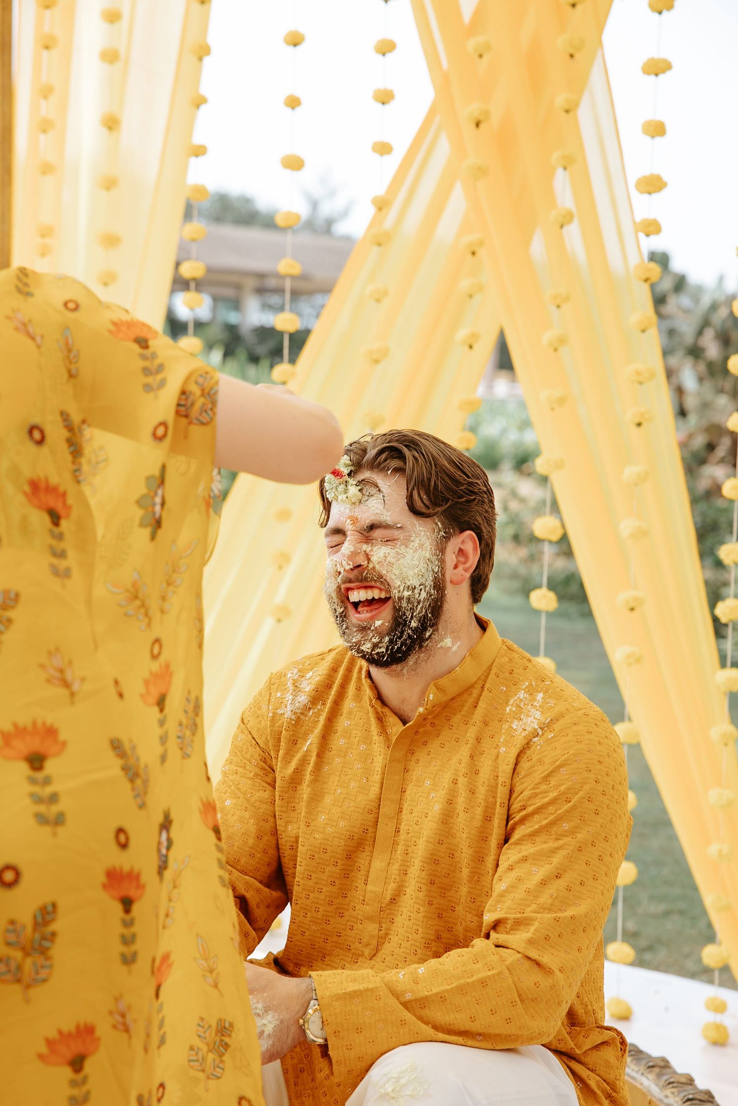 Laughing groom in mustard kurta gets haldi applied on face during Indian wedding ceremony under yellow decor.