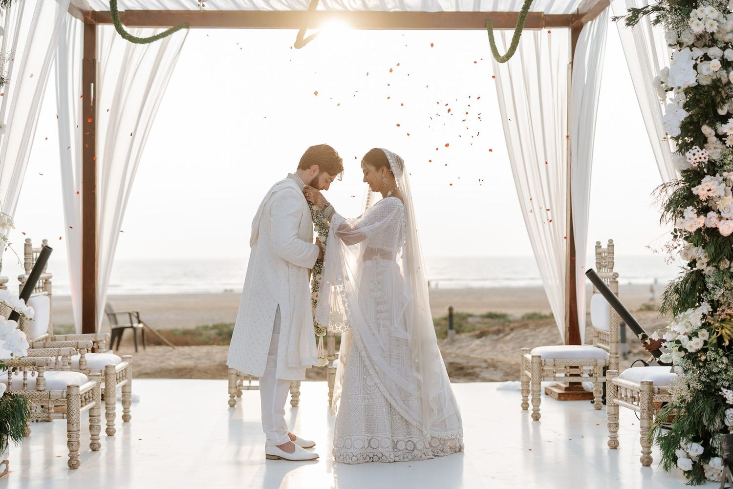 Couple exchanging vows at elegant beach wedding ceremony under white floral arch with ocean backdrop.