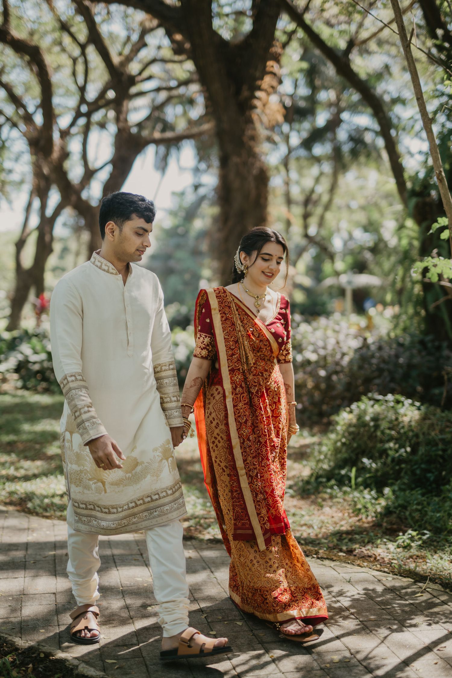 A couple in traditional South Asian wedding attire walk together on a garden path under lush trees.