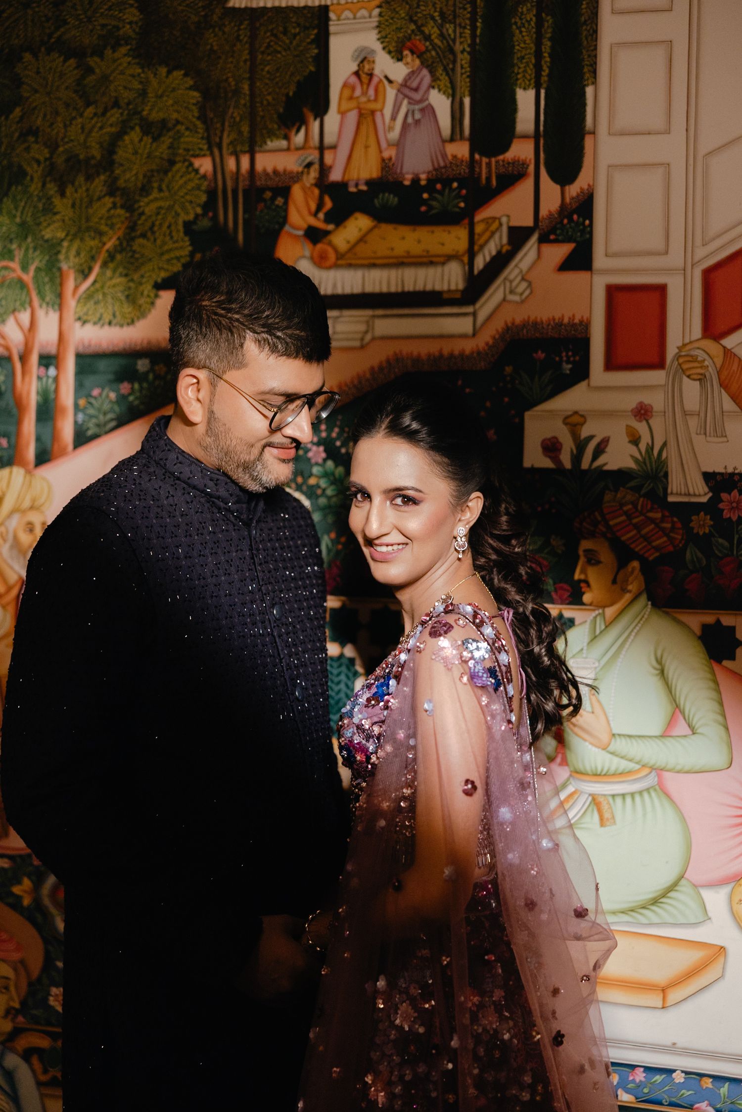 A couple shares romantic moments in traditional Indian attire at an ornately decorated venue with colorful wall murals.