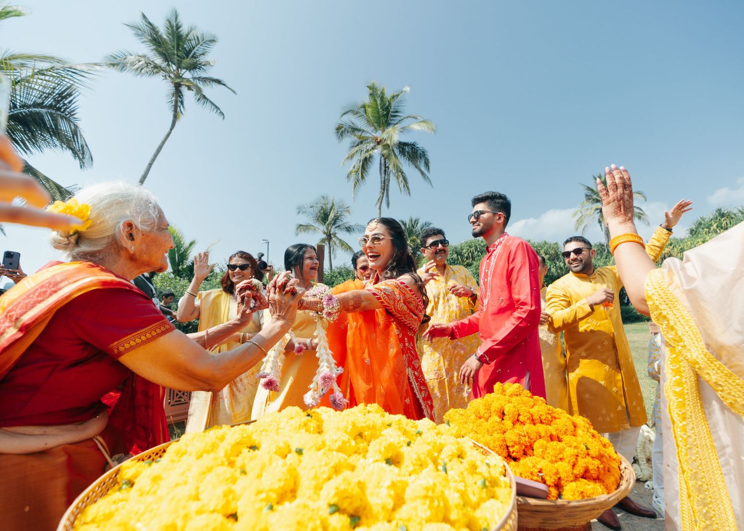 Festive group celebrates with yellow marigold flowers and traditional attire during an outdoor ceremony with palm trees.