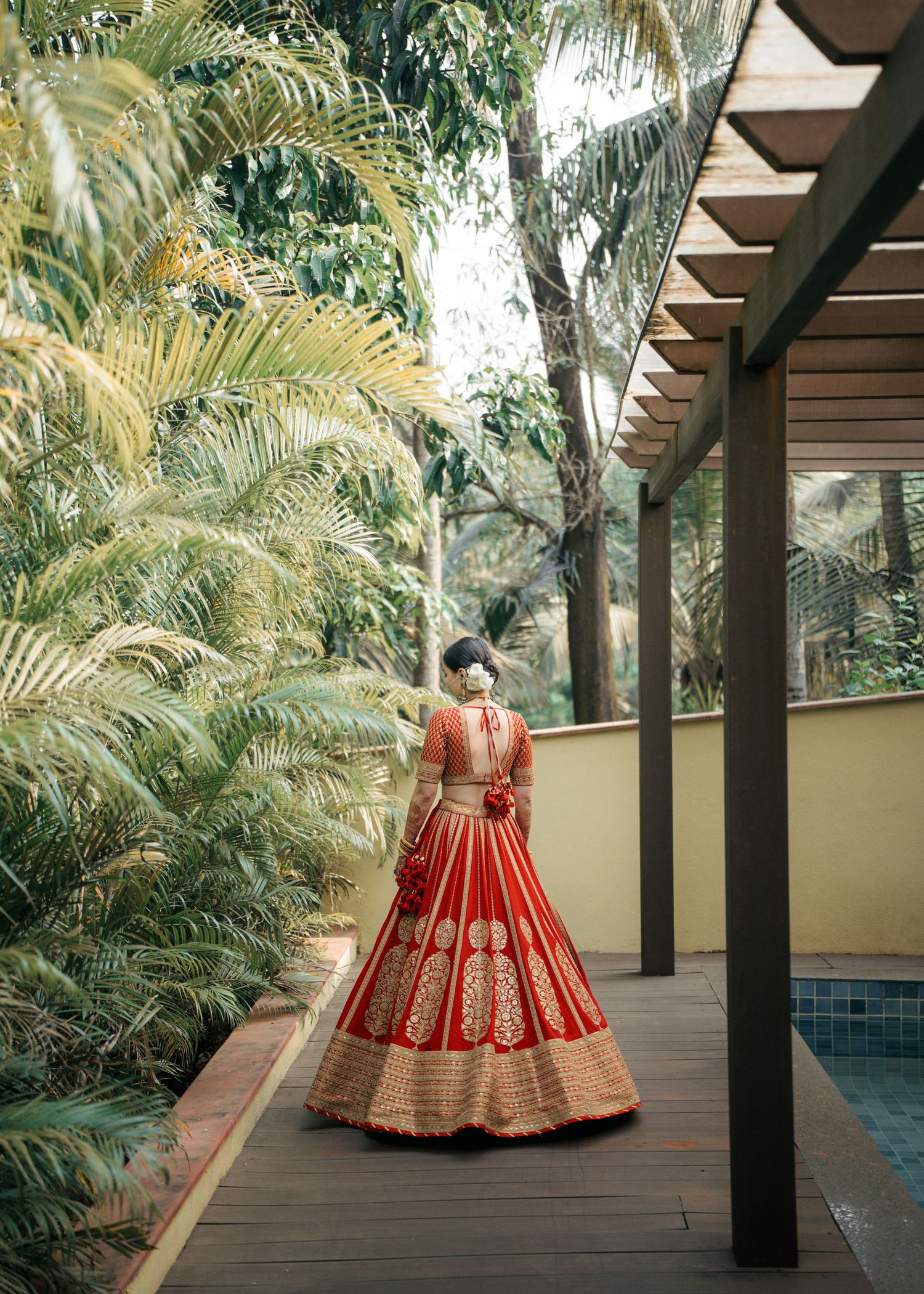 A red and gold traditional Indian lehenga dress photographed from behind on a wooden deck surrounded by tropical palm trees.