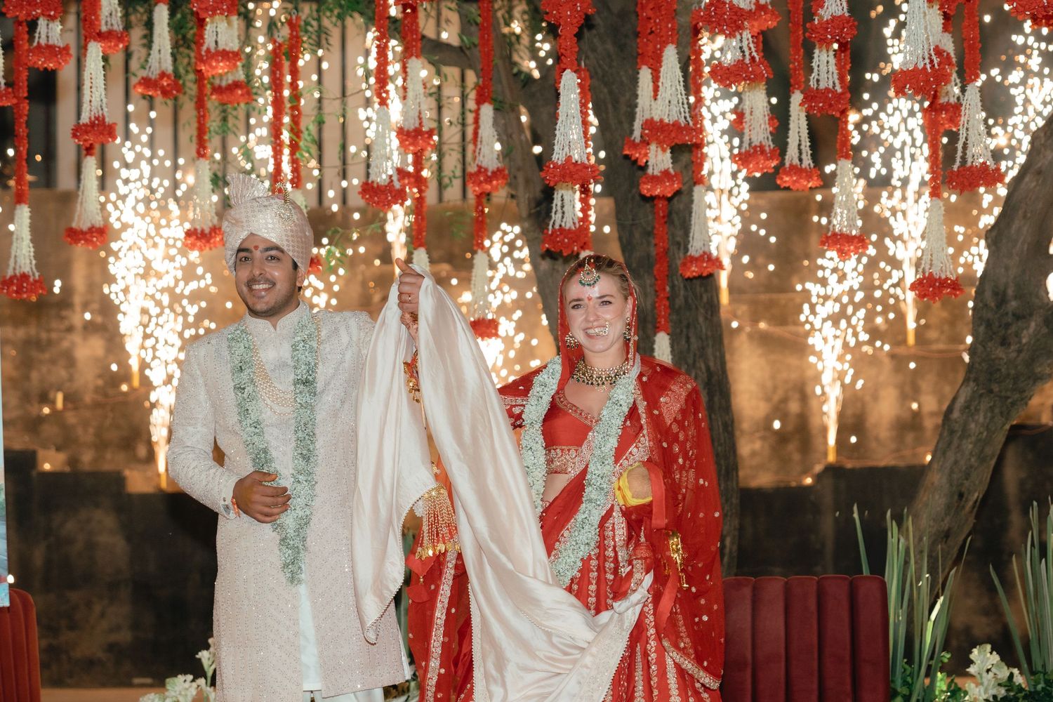 Newlyweds in traditional Indian wedding attire standing under red and white floral decorations.