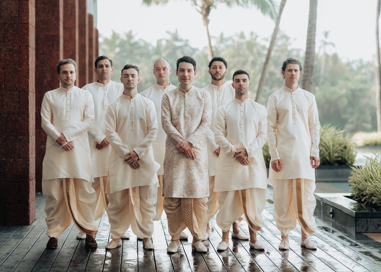 Groomsmen in matching ivory traditional outfits posing together on wooden deck.