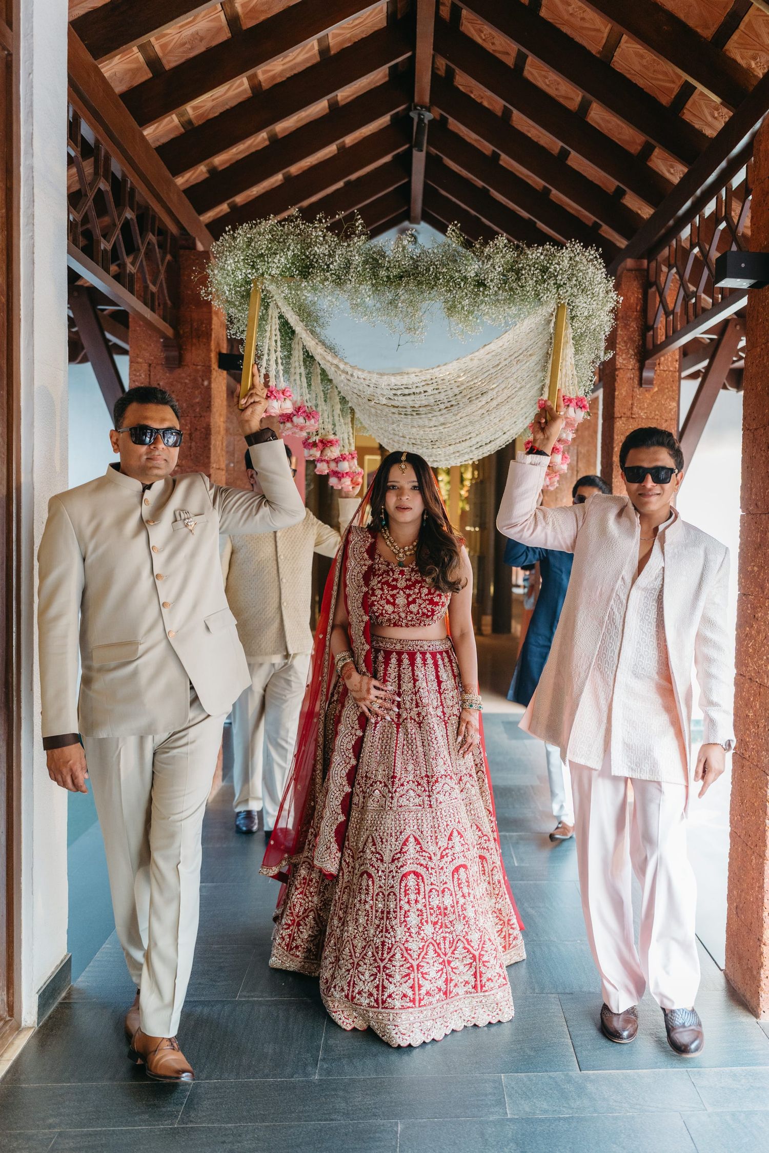 Bride in red lehenga walks under floral canopy escorted by men in white suits at Indian wedding ceremony.