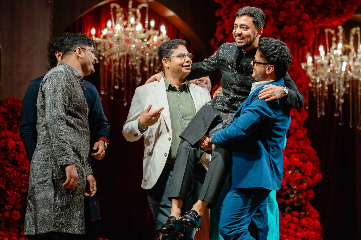 Men in festive attire laughing and embracing at a glamorous red-themed celebration with chandelier backdrop.
