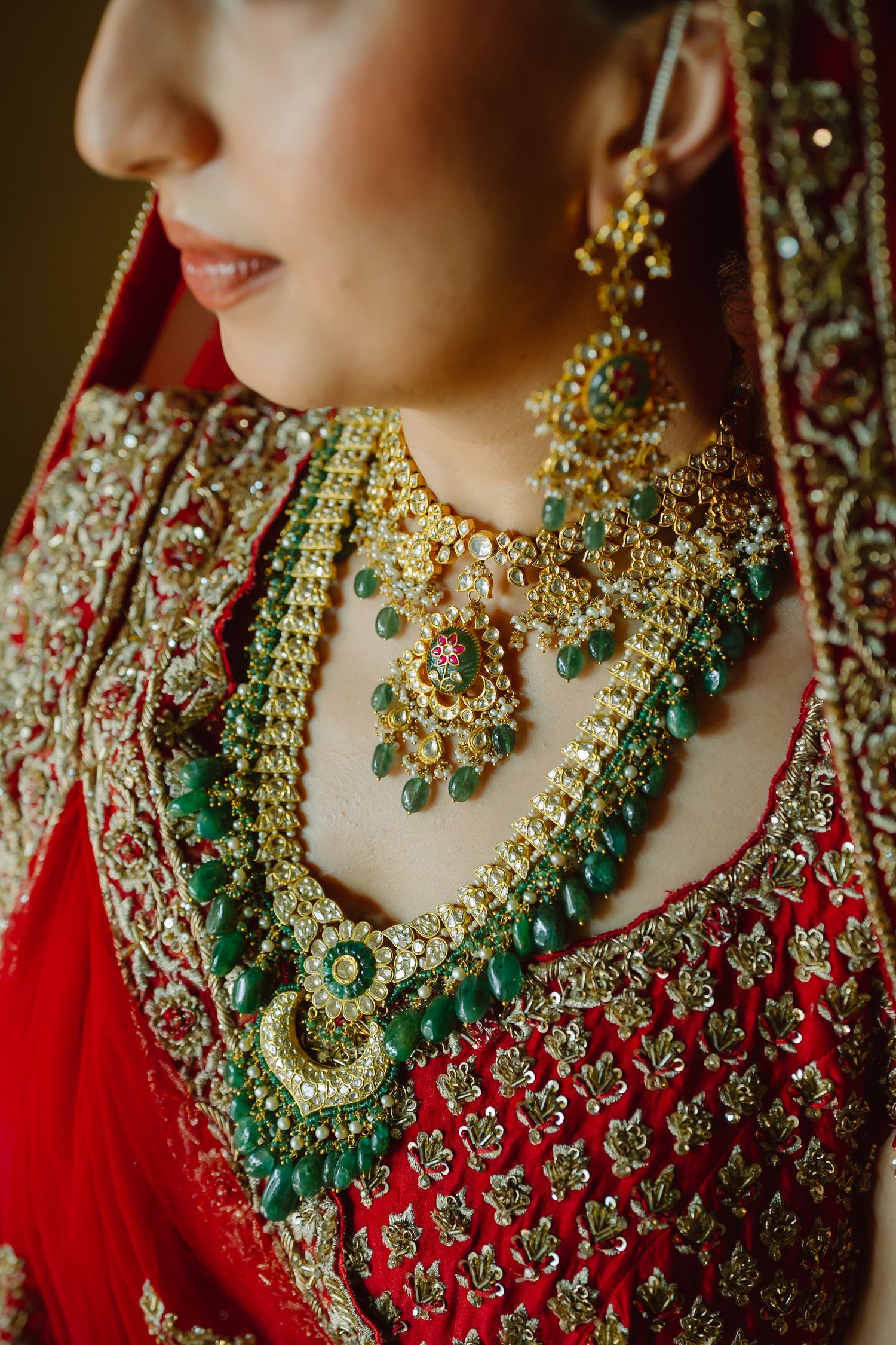 Indian bride in red lehenga wearing layered gold and emerald green bridal jewelry including necklace and earrings.