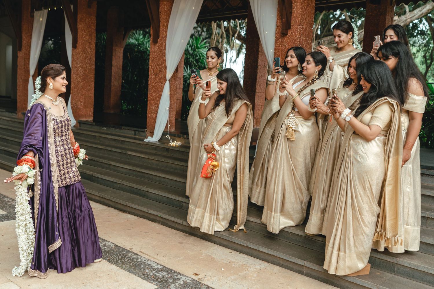 Bride in purple sharara stands before bridesmaids in gold sarees at an elegant Indian wedding ceremony.