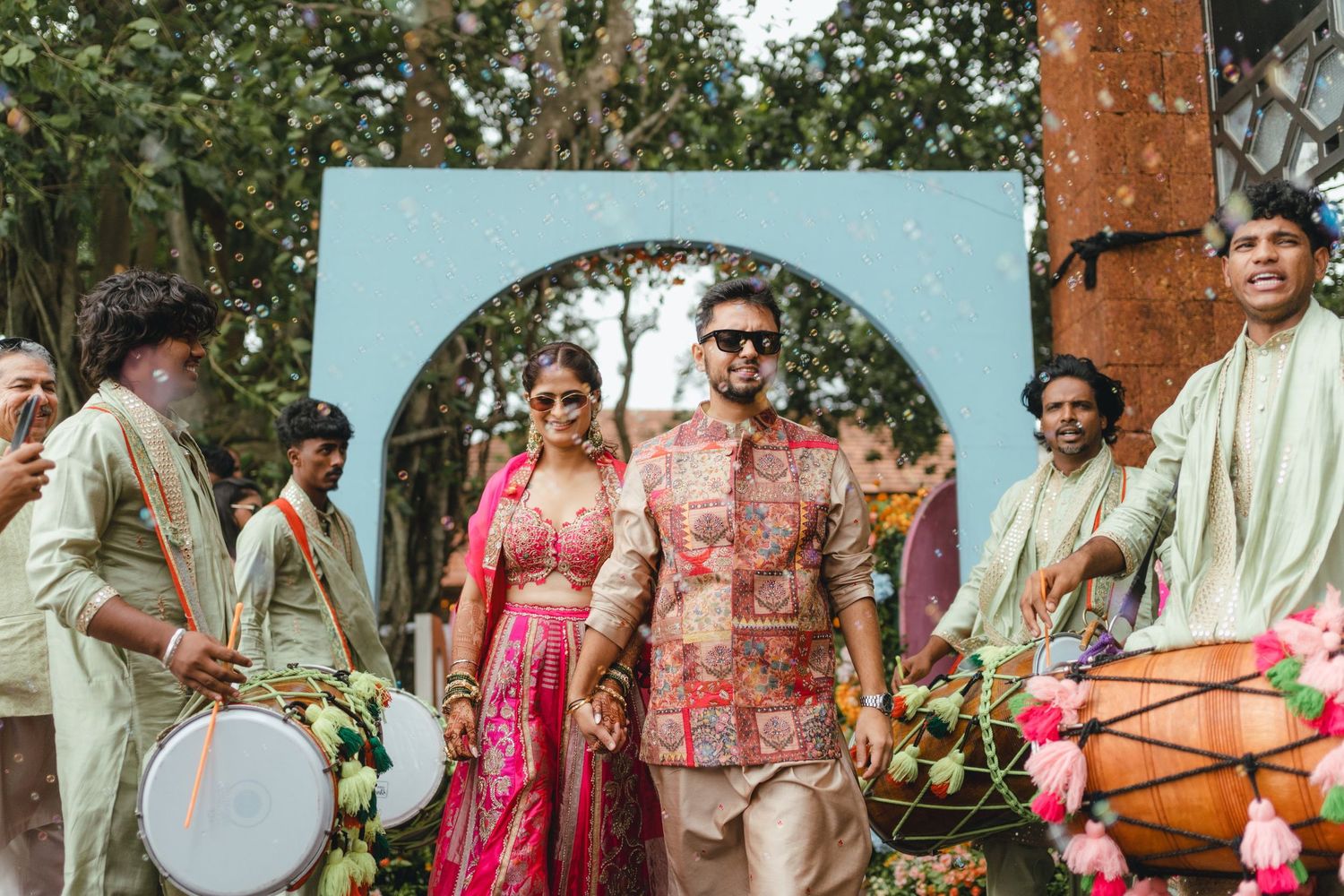 Indian bride and groom walking together at traditional wedding ceremony with drummers and floral decorations outdoors.