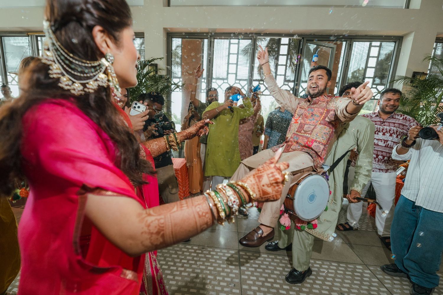 Vibrant Indian wedding celebration with guests dancing, drummer playing dhol, bride in red lehenga with mehndi, joyful festive atmosphere.
