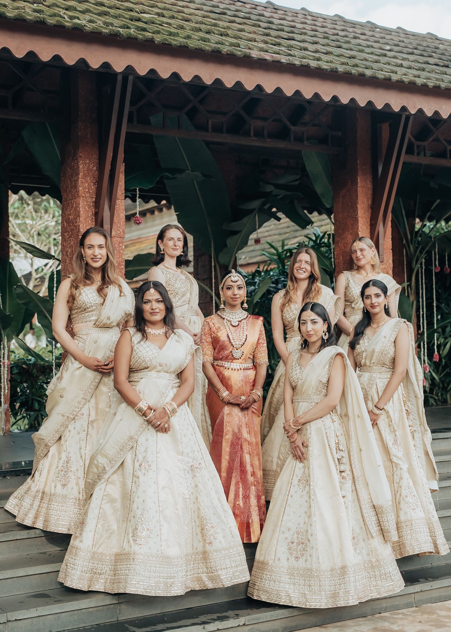 A group of women in elegant traditional Indian wedding attire pose together in cream and gold lehengas at an outdoor venue.